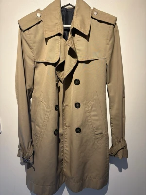 Beige klassisk trenchcoat med bälte - Stilren beige trenchcoat med dubbelknäppning, axelklaffar och bälte i midjan. Jackan har två snedställda fickor framtill och justerbara ärmslut. Perfekt för dig som vill ha en tidlös och trendig look.