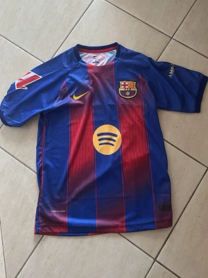 FC Barcelona matchtröja Nike - Säljer en officiell FC Barcelona matchtröja från Nike med Lamine Yamal och nummer 10 på ryggen. Tröjan är blå och vinröd med gula detaljer, klubbmärke och sponsorloggor. Tillverkad i lätt och ventilerande material, perfekt för fotboll eller träning.