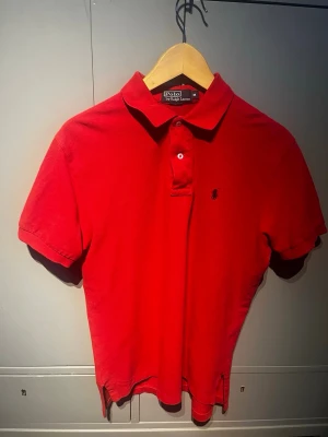 Röd pikétröja från Ralph Lauren - Klassisk röd pikétröja från Polo Ralph Lauren med korta ärmar, krage och knappar framtill. Tröjan är i bomull och har den ikoniska broderade loggan på bröstet. Perfekt för en clean och stilren look. Storlek M