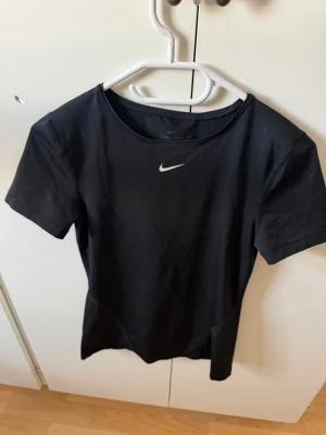 Svart Nike t-shirt med logga - Säljer en svart t-shirt från Nike med klassisk vit logga på bröstet. T-shirten har rund hals och korta ärmar, perfekt för dig som gillar sportiga och stilrena plagg. Materialet är polyester och modellen är slim fit.