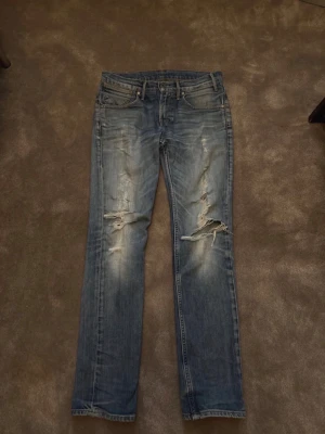 Blå slitna jeans från Levi's - Säljer ett par blå jeans från Levi's med snygga slitningar och hål på benen. Jeansen har klassisk femficksdesign, raka ben och tydliga slitdetaljer framtill. Perfekta för en avslappnad och trendig look.