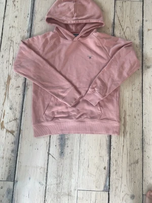 Rosa hoodie från Gant - Säljer en snygg rosa hoodie från Gant med liten broderad logga på bröstet. Hoodien har klassisk känguruficka, ribbade muddar och huva. Perfekt för en chill och avslappnad stil. Tillverkad i mjuk bomullsblandning.