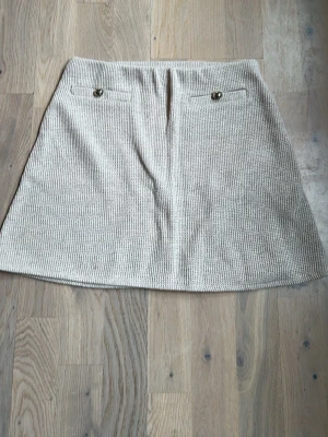 Beige stickad kjol från H&M - Beige stickad kjol från H&M med två dekorativa fickor framtill och silvriga knappar. Kjolen har en rak, kort passform och är gjord i ett mjukt stickat material som känns både trendigt och bekvämt.