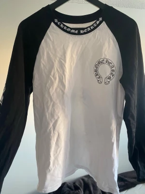 Chrome Hearts longsleeve - Säljer en långärmad topp från Chrome Hearts med svartvita kontraster och tryck både fram och bak. Tröjan har svart ärmar och vit kropp, samt tydliga logotyper och textdetaljer. Perfekt för dig som gillar streetwear och vill sticka ut. Riktigt fint skick! 👌
