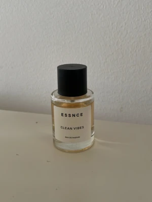 ESSNCE Clean Vibes Eau de Parfum - Parfymflaska från ESSNCE med doften Clean Vibes. Flaskan är cylindrisk i genomskinligt glas med svart rund kork och vit etikett med svart text. Modern och stilren design som passar perfekt på parfymhyllan.