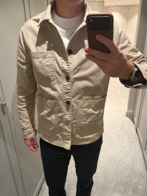 Beige overshirt i bomull från J.Lindeberg - Stilren beige overshirt med klassisk krage och tre stora fickor framtill. Jackan har knäppning med stora knappar och är tillverkad i ett mjukt bomullstyg. Perfekt för lager-på-lager och enkel att matcha med olika outfits.