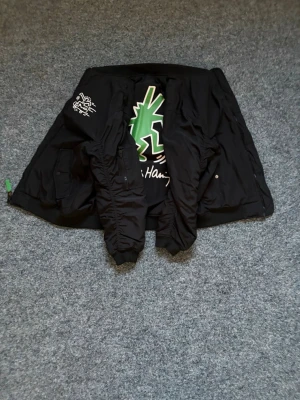Svart bomberjacka Keith Haring print - Snygg svart bomberjacka från Alpha Industries med Keith Haring-inspirerat tryck på bröstet och stor grön figur på ryggen. Jackan har orange innerfoder, ribbade muddar och fickor med lock samt ärmficka med dragkedja. Perfekt för dig som gillar streetwear och unika prints.