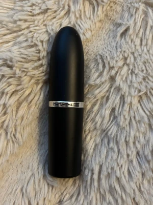MAC Forever Curious matt läppstift - MAC Forever Curious är ett matt läppstift i klassisk svart hylsa med silverdetalj. Färgen är neutral och passar till många olika looks. Hylsan har den ikoniska MAC-loggan och är tillverkad i plast med metallisk ring.
