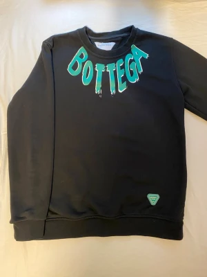 Svart sweatshirt från Bottega Veneta - Cool svart sweatshirt från Bottega Veneta med stort grönt och vitt tryck på både fram- och baksidan. Tröjan har rund hals, långa ärmar och ribbade muddar. 