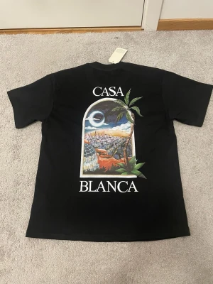Svart Casa Blanca t-shirt med tryck - Svart t-shirt från Casa Blanca med färgglatt tryck av stadsmotiv och palmer på både bröstet och ryggen. Klassisk passform med rund hals och korta ärmar. Perfekt för dig som vill sticka ut med en unik design.