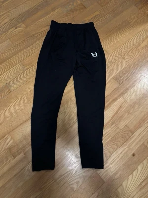 Svarta mjukisbyxor från Under Armour - Svarta mjukisbyxor från Under Armour med elastisk midja och smal passform. Byxorna har en vit logga på vänster lår och är tillverkade i ett mjukt och stretchigt material som passar perfekt för träning eller chill. Klassisk look med diskreta sömmar.