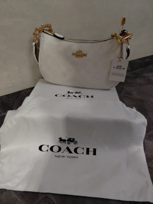 Vit axelremsväska från Coach - Snygg vit axelremsväska från Coach med guldfärgade detaljer och logotyp framtill. Väskan har ett diskret mönster i materialet och levereras med dustbag och tag. Perfekt för dig som vill ha en stilren och lyxig accessoar.