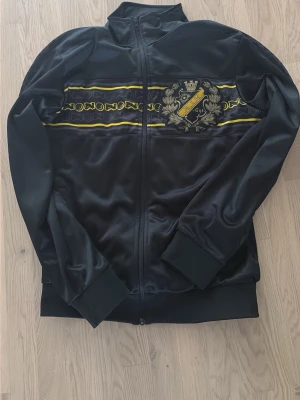 Svart AIK ziptröja med emblem - Svart ziptröja från AIK med klubbens emblem och årtal broderat på bröstet. Tröjan har gula och vita detaljer över bröstet, hög krage och muddar vid ärmslut och midja. Tillverkad i glansigt syntetmaterial, perfekt för dig som vill visa ditt AIK-stöd.