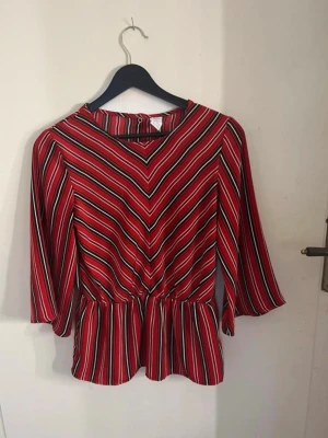 Röd randig blus med trekvartsärm - Snygg röd blus med svarta och vita diagonala ränder framtill och vertikala ränder baktill. Blusen har trekvartsärmar, rund halsringning och markerad midja med volang nedtill. Tillverkad i ett lätt och mjukt tyg, perfekt för en trendig look. Fin röd blus. Bra passform och bra i storleken men passar även dam i S eftersom den är lite stor. Super fin på och sitter bra, skönt tyg. Storlek 158/S. Den är nästan aldrig använd! 