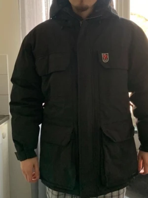 Svart parkas från Fjällräven - Svart parkas från Fjällräven med klassisk logga på bröstet. Väldigt bra skick förutom som man ser på sista bilden då där man spänner är lite sönder men går att fixa rätt lätt tror jag men syns inte alls när man har på sig den. Jag är 186 lång så den är en stor Small.               Nypris runt 4000