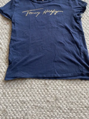  Tommy Hilfiger t-shirt  - Snygg marinblå t-shirt från Tommy Hilfiger med guldfärgad logga i skrivstil på bröstet. Klassisk passform och rund halsringning. Perfekt till jeans eller shorts för en avslappnad look. Skriv för fler bilder🤍🤍 