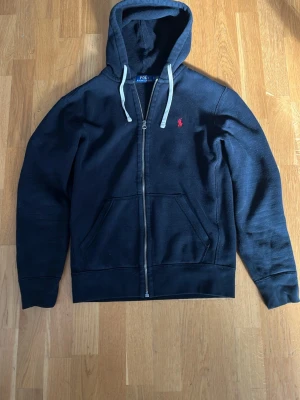 Svart hoodie från Polo Ralph Lauren - Svart hoodie med dragkedja från Polo Ralph Lauren. Den har en huva med vita snören, två fickor framtill och ett litet rött logotryck på bröstet. Perfekt för en avslappnad och cool look. Materialet känns mjukt och bekvämt.