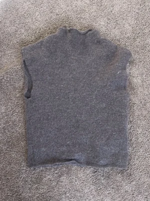 Mörkgrå stickad väst med hög krage - En enkel och stilren mörkgrå stickad väst med hög krage. Perfekt att slänga över en skjorta eller tröja för en chill lager-på-lager-look. Västen har en rak passform och är gjord i mjukt material som känns skönt mot huden.