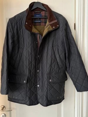 Svart quiltad jacka från Gant XL - Svart quiltad jacka från Gant. Jackan har rutigt innerfoder i grönt, brunt och rött samt bruna manchesterdetaljer vid kragen. Två stora fickor framtill med tryckknappar och dragkedja samt knappstängning. Jättefräsch och snygg jacka till killar!