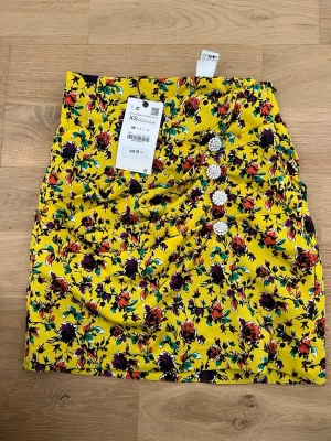Gul blommig kortkjol med knappar - Färgstark gul kortkjol från ZARA med blommigt mönster i lila, rött och grönt. Kjolen har dekorativa glittriga knappar framtill och en figurnära passform. Perfekt för dig som vill sticka ut med en unik och trendig look. Den är helt ny 