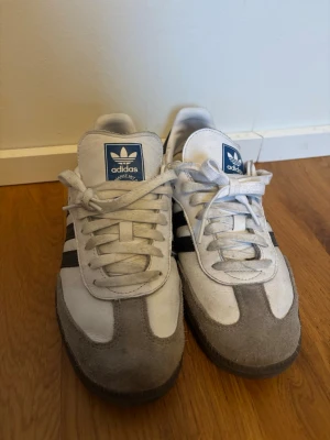 Adidas Samba vita och grå sneakers - Klassiska Adidas Samba sneakers i vitt läder med grå mockadetaljer och svarta ränder på sidorna. Skorna har blå Adidas-logga på plösen och mörk sula. Perfekt retrostil med låg profil och tidlös design. Skorna är i princip helt nya bara använda ett go tal gånger. Priset är ej hugget i sten😊