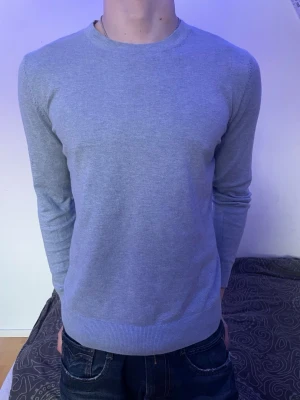 Ljusgrå basic tröja med rund hals - En stilren ljusgrå tröja med rund halsringning och långa ärmar. Tröjan har en smal passform och ribbade muddar vid ärmslut och nederkant. Perfekt att matcha med jeans för en enkel och clean look.