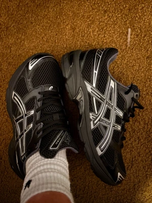 Asics Gel-1130 Black Silver - Snygga svarta Asics sneakers med silvriga detaljer och klassisk logga på sidorna. Ovandelen är i mesh och syntet för bra andningsförmåga och komfort. Skorna har en sportig vibe och passar perfekt till streetwear eller casual outfits. Passar strl 43.