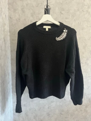 Svart stickad tröja med brosch - Svart stickad tröja med rund halsringning och långa ärmar. På vänster axel sitter en dekorativ brosch i form av ett silverfärgat löv med små stenar🎀🎀