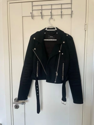 Svart  från Pull&Bear  - Snygg svart jacka från Pull&Bear med silvriga dragkedjor, axelklaffar och bälte i midjan. Jackan har två framfickor med dragkedja och coola detaljer på ärmarna. Perfekt för en edgy look.