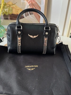 Svart handväska Zadig & Voltaire - Säljer en svart handväska från Zadig & Voltaire i strukturerat läder. Väskan har silverfärgade detaljer, dubbla dragkedjor framtill och ett emblem med vingar. Den har både handtag och avtagbar axelrem för olika stilar.
