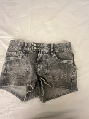 Grå denimshorts med broderade fickor - Snygga grå denimshorts med tvättad look och dubbla knappar fram. Shortsen har stora framfickor och bakfickor med broderade hjärtan och snirkliga detaljer. Perfekta för sommaren och har bälteshällor för extra stil.