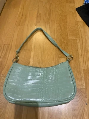 Ljusgrön axelväska i krokopräglat skinn - Snygg ljusgrön handväska med axelrem och krokodilpräglat mönster. Väskan har guldfärgade metalldetaljer och dragkedja upptill. Insidan har ett fack med dragkedja och ett öppet fack för småsaker. Perfekt för dig som gillar trendiga accessoarer.