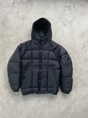 Svart pufferjacka med huva C.P. Company - Svart pufferjacka från C.P. Company med huva och dragkedja framtill. Jackan har två snedställda fickor med dragkedja och en ikonisk linsdetalj på vänster ärm. Quiltad design och fyllig vaddering för extra värme.