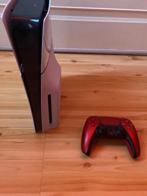 PlayStation 5 - PlayStation 5 konsol i vit och svart med futuristisk design och en röd DualSense trådlös handkontroll. Konsolen har diskfack och är tillverkad i plast med matta och blanka ytor. Perfekt för gaming på nästa nivå.