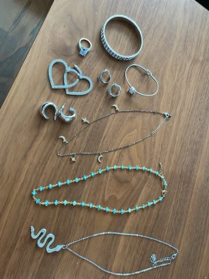 Mix av silversmycken och halsband - Säljer ett set med silversmycken: armband, ringar, örhängen (hjärtan, hoops, månar), samt tre halsband – ett med stjärnor och månar, ett med blåvita pärlor och ett med en glittrig orm. Flera smycken har stenar och gnistrande detaljer. Perfekt för dig som gillar att mixa stilar. Alla halsband är i ett bra skick, örhängen är oanvända/nytt skick och armband är i mycket bra skick allt kommer från ur&penn och glitter. Vill du ha en viss del av smyckena så skrivnoch vi löser det!