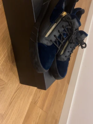 Giuseppe zanotti skor med guldig dragkedja  - Snygga sneakers i mörkblå sammet och svart läder från Giuseppe Zanotti. Skorna har guldfärgade dragkedjor på sidorna och svarta snören. Tjock sula och cool design som sticker ut. Perfekta för dig som vill ha något unikt och trendigt.