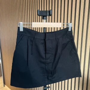 Svart kostymkjol från H&M - Stilren svart kortkjol med fickor fram och bälteshällor i midjan. Kjolen har en klassisk rak passform och är perfekt att matcha med både sneakers och boots. Materialet känns som bomull eller polyester och kjolen har diskreta sömdetaljer framtill.