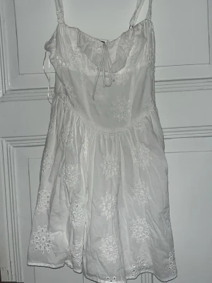 Vit broderad sommarklänning - Söt vit kortklänning med broderade blommor och smala axelband. Klänningen har knytning framtill och är gjord i ett lätt bomullstyg, perfekt för varma dagar. Den har en luftig passform och fina detaljer som ger en romantisk känsla. Fårn Gina, knappt använd och är för liten nu. Köptes för 600kr. Jätte bra skick utan defekter. 