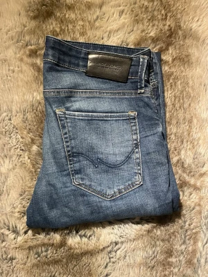 Blå slim fit jeans från Jack & Jones W30L30 - Snygga blå slim fit jeans från Jack & Jones med klassisk femficksdesign och diskreta slitningar framtill. Jeansen har en mörkare tvätt och är tillverkade i bomull med lite stretch för extra komfort. Perfekta för en avslappnad och trendig look.