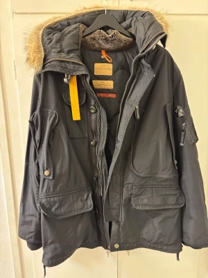 Svart jacka från Parajumpers - Säljer en svart jacka från Parajumpers med avtagbar fuskpäls på huvan, flera stora fickor och gul detalj vid halsen. Jackan har quiltat innerfoder och dragkedja med knappar. Perfekt för kalla dagar och riktigt snygg streetstil.