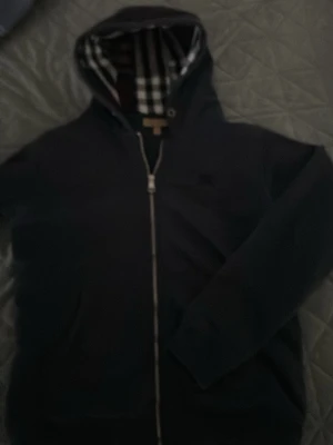 Svart hoodie med Burberry-mönstrad luva - Snygg svart hoodie med dragkedja från Burberry. Insidan av luvan har det klassiska rutmönstret i svart, vitt och rött. Hoodien har långa ärmar och ribbade muddar. Perfekt för dig som vill ha en stilren och exklusiv look.