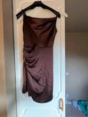 Brun asymmetrisk kort klänning - Säljer en brun, kort klänning med asymmetrisk axel och draperad detalj på sidan. Klänningen har smala axelband och är tillverkad i ett glansigt material som ger en elegant look. Dragkedja i ryggen för enkel påklädning.