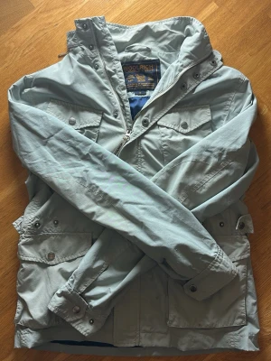 Field jacket från Woolrich - Snygg ljusgrå Field jacket från Woolrich med flera fickor och detaljerade knappar med logga. Jackan har hög krage och justerbara ärmslut. Tillverkad i lätt och slitstarkt material, perfekt för blåsiga dagar. Passar dig som är runt 180 cm lång,  vid övriga frågor är det bara att höra av sig🤩