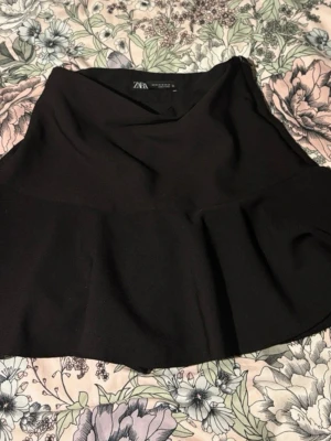 Svart kortkjol från Zara - Kort svart kjol från Zara med lätt utsvängd form. Kjolen är enkel och stilren, tillverkad i ett mjukt syntetmaterial som ger en snygg siluett. Perfekt att styla med en crop top eller oversized tröja. Kosymbyxor material hade ett band runt frånbörjan som jag klippte bort  