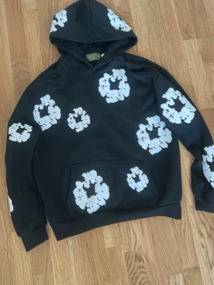 Svart hoodie med vita blommönster - Cool svart hoodie med stora vita blommönster över hela tröjan, inklusive huvan. Hoodien har en klassisk känguruficka framtill och ribbade muddar vid ärmslut och nederkant. Perfekt för dig som vill sticka ut med en unik och trendig stil.