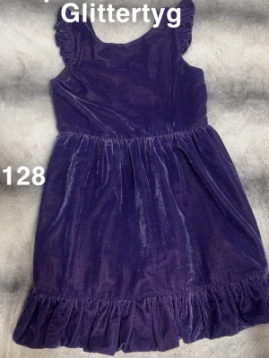 Lila glittrig klänning med volang - Supersöt lila klänning i glittrigt tyg med volangkant nedtill och små volanger vid axlarna. Klänningen har en rund halsringning och är ärmlös, perfekt för dig som vill sticka ut lite extra.🌸 Sparsamt använd. Glittertyg. Storlek 128
