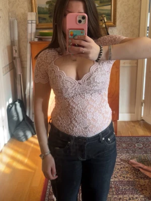 Ljusrosa spetsbody med v-ringning  - Supersnygg ljusrosa body i spets med v-ringad hals och korta ärmar. Kroppsnära passform och fina spetsdetaljer över hela toppen. Perfekt att styla med jeans eller kjol för en trendig look.