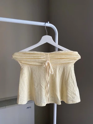 Gul offshoulder spets topp med knyt - Söt ljusgul offshoulder topp med spetsdetaljer och knyt framtill. Toppen har en luftig passform och är perfekt för varma dagar. Materialet är lätt och har ett diskret hålmönster som ger en romantisk känsla.