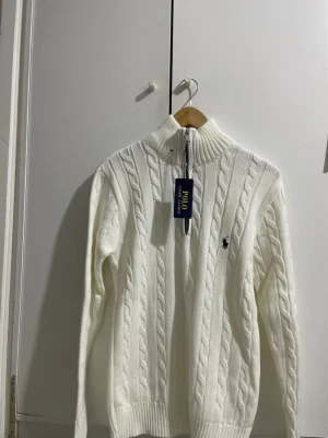 Vit kabelstickad tröja Polo Ralph Lauren - Kabelstickad vit tröja från Polo Ralph Lauren med halv dragkedja och hög krage. Tröjan har det klassiska broderade logotypen på bröstet och ribbade muddar vid ärmslut och nederkant. Perfekt för en stilren och klassisk look.