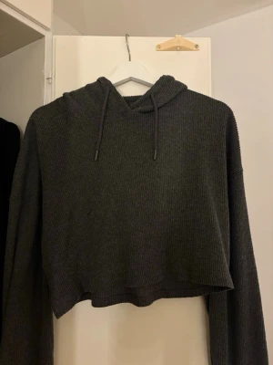 Grå cropped hoodie från Sisters Point - Mörkgrå ribbstickad cropped hoodie från Sisters Point. Hoodien har långa ärmar, dragsko i huvan och är tillverkad i ett mjukt, stickat material. Perfekt för en avslappnad och trendig look.